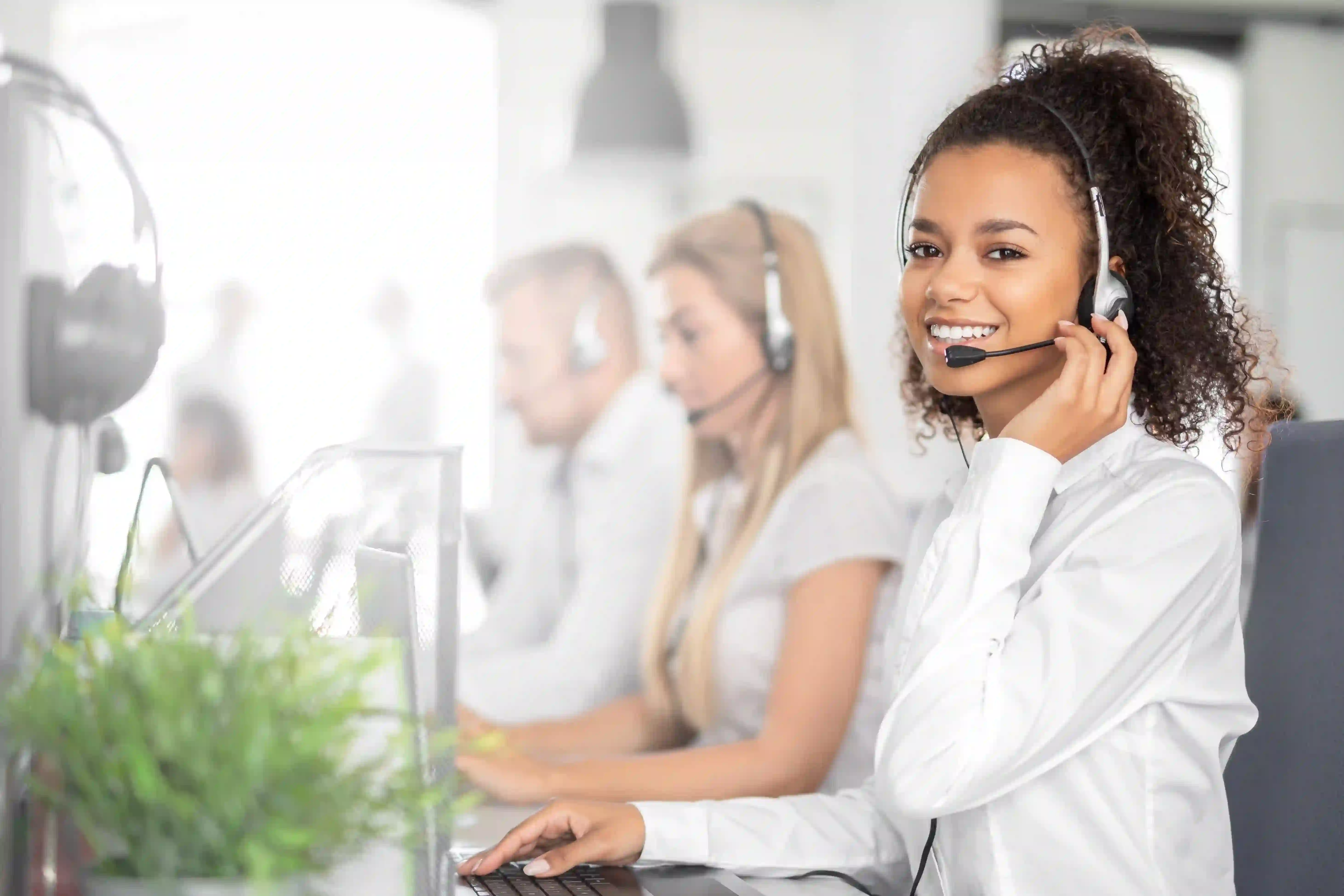 En smilende kvinde med headset yder professionel telefonpasning i et moderne callcenter. Hun sidder foran en computer og besvarer opkald på vegne af kunder, mens to kollegaer i baggrunden også arbejder koncentreret. Miljøet er lyst og organiseret, hvilket afspejler en effektiv og venlig kundeservice.