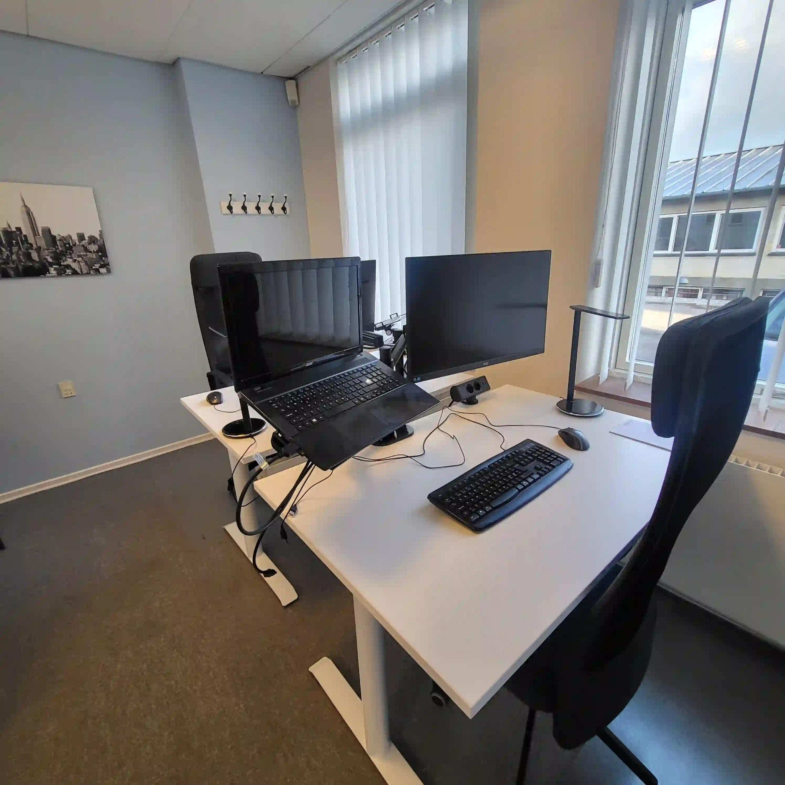 Et moderne og lyst kontor med hæve-/sænkebord, ergonomisk kontorstol, stor computerskærm og vindue med masser af naturligt lys. Kontoret kan lejes på timebasis hos Flexum i Kongens Lyngby.