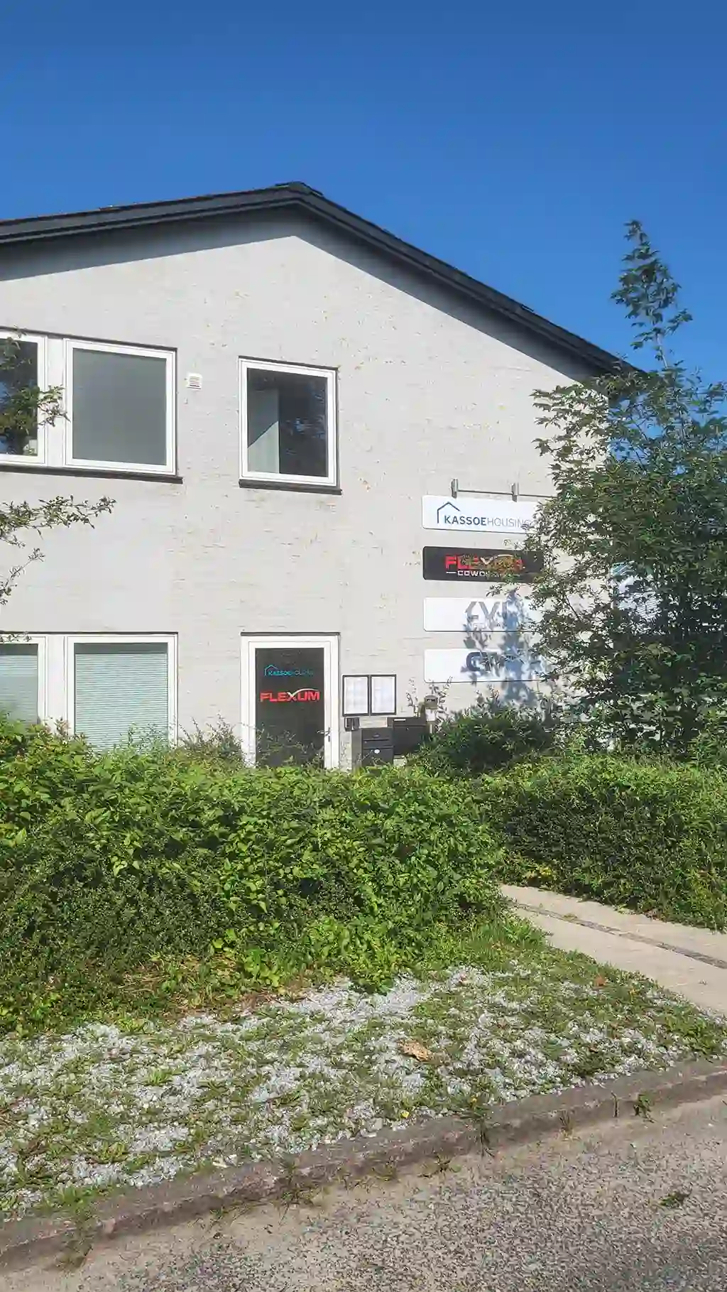 Facade af Flexum Coworking bygning på en solrig dag, beliggende på Maglebjergvej 6. Den lysegrå to-etagers kontorbygning har store vinduer og tydelig skiltning med Flexum Coworking logo ved indgangen. Omgivelserne er grønne og indrammet af træer og buske.