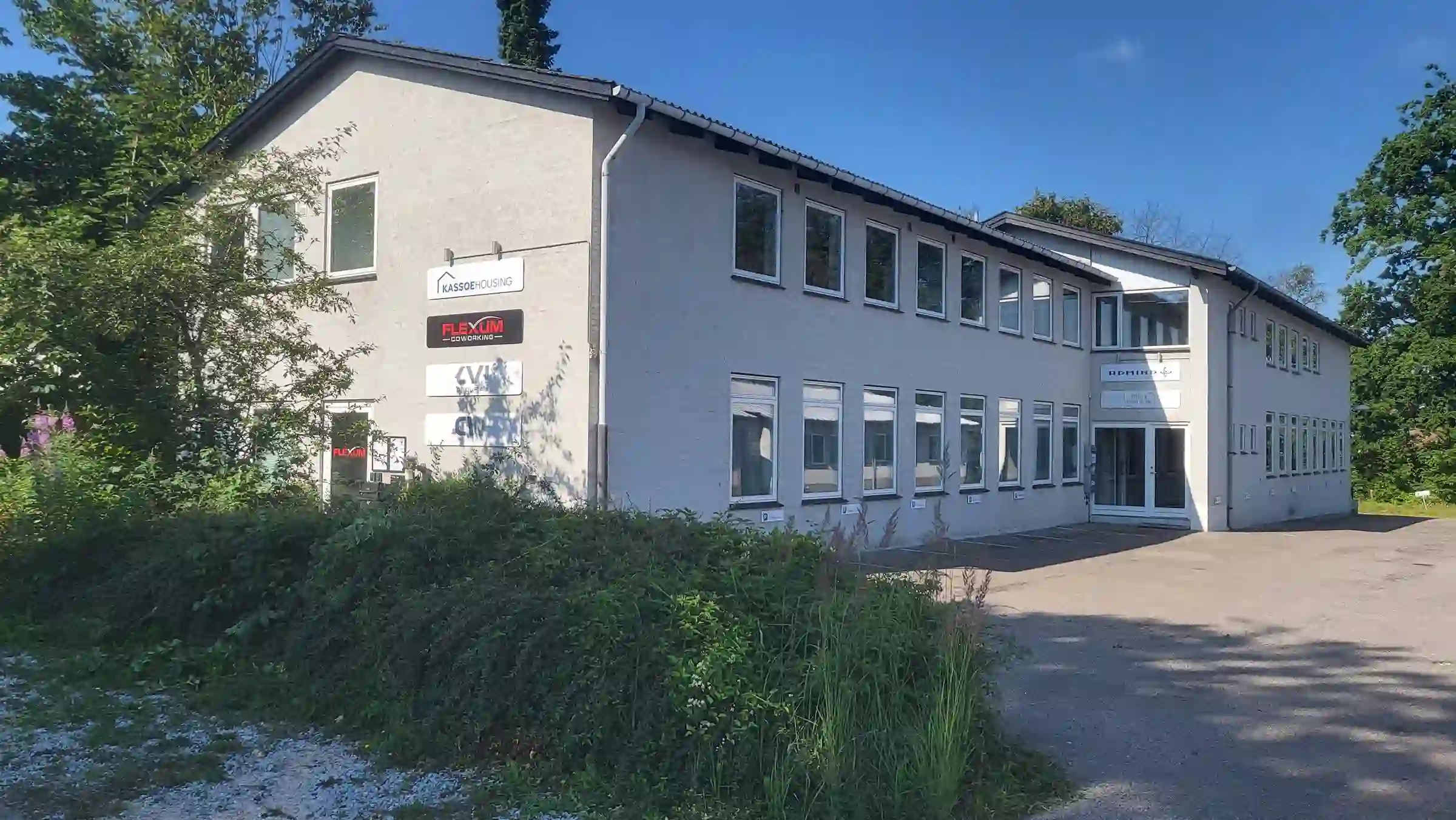 Facade af Flexum Coworking bygning på en solrig dag, beliggende på Maglebjergvej 6. Den lysegrå to-etagers kontorbygning har store vinduer og tydelig skiltning med Flexum Coworking logo ved indgangen. Omgivelserne er grønne og indrammet af træer og buske.