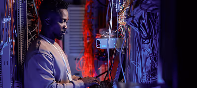 Un técnico de TI, un hombre negro, trabaja en equipos en medio de una densa red de cables y servidores en un centro de datos, iluminado por una luz azul y violeta.