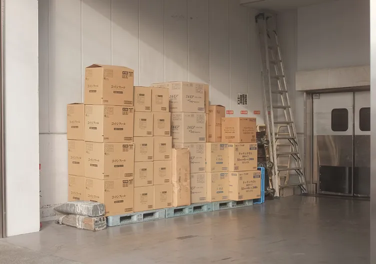 A pallet stacked high with cardboard boxes sits on the floor of a warehouse next to a tall ladder and a large metal door