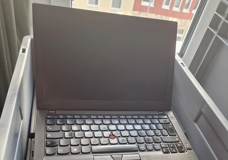 A black Lenovo ThinkPad laptop with a red TrackPoint button on the keyboard is sitting open inside a large grey plastic bin