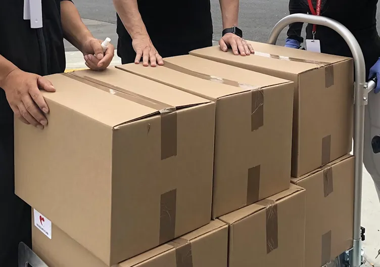 Three people stand around a dolly stacked with cardboard boxes. One person appears to be spraying sanitizer on their hands
