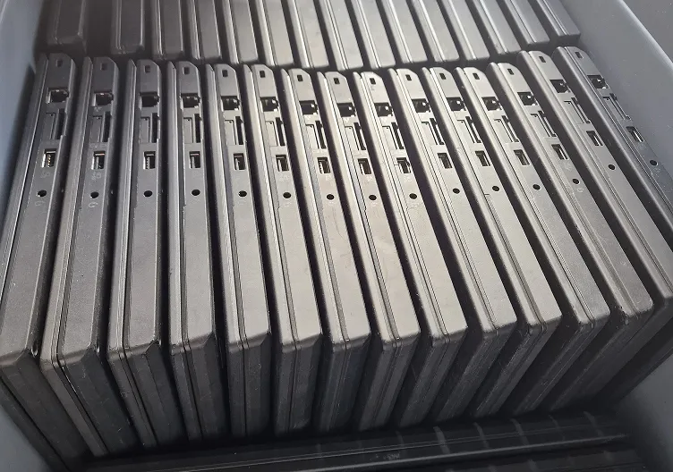 A top-down view of numerous closed black laptops packed vertically side-by-side in a container, showing their hinge and port sides