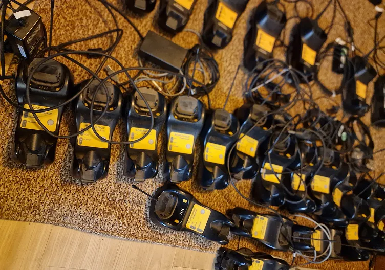 Numerous black handheld electronic devices with yellow labels and attached cables are laid out on a carpeted floor