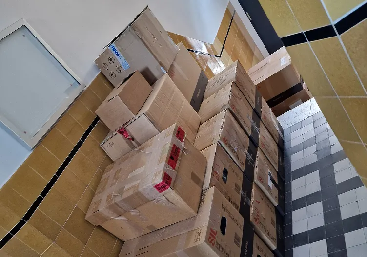 A hallway with yellow and white tiled walls and a black and white tiled floor, filled with a large, disorganized stack of cardboard boxes of various sizes