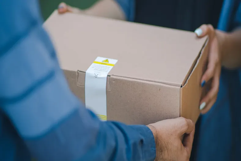 A close-up of a cardboard package being handed from one person, whose blue-striped sleeve is visible, to another person