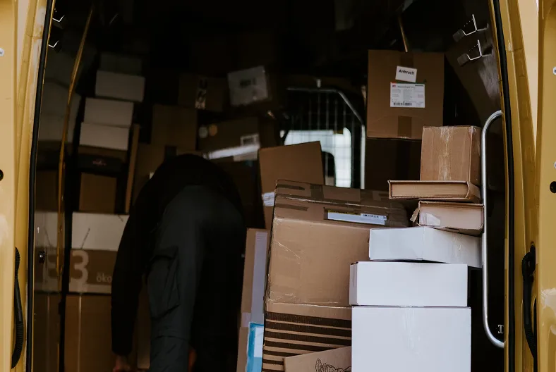 The back of a person in dark clothing is visible as they reach into a yellow delivery van packed with cardboard boxes