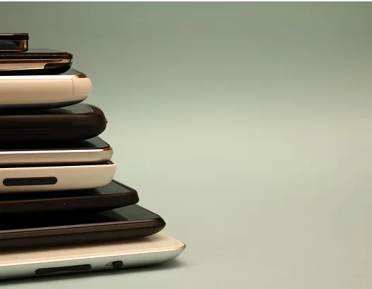 A close-up, side-view of a stack of smartphones in different colors, including white, silver, and black, against a plain light-colored background