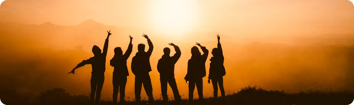The silhouette of a group of six people standing on a hill with their arms raised in celebration against a golden sunset