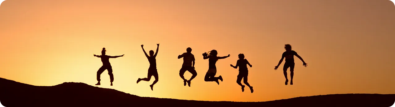 The silhouette of six people in mid-air, jumping enthusiastically on a hill against a clear orange sky at sunset.