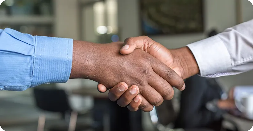 A close-up of two men shaking hands in a professional setting. One man is wearing a blue shirt, and the other is wearing a white shirt, with a blurred office in the background.