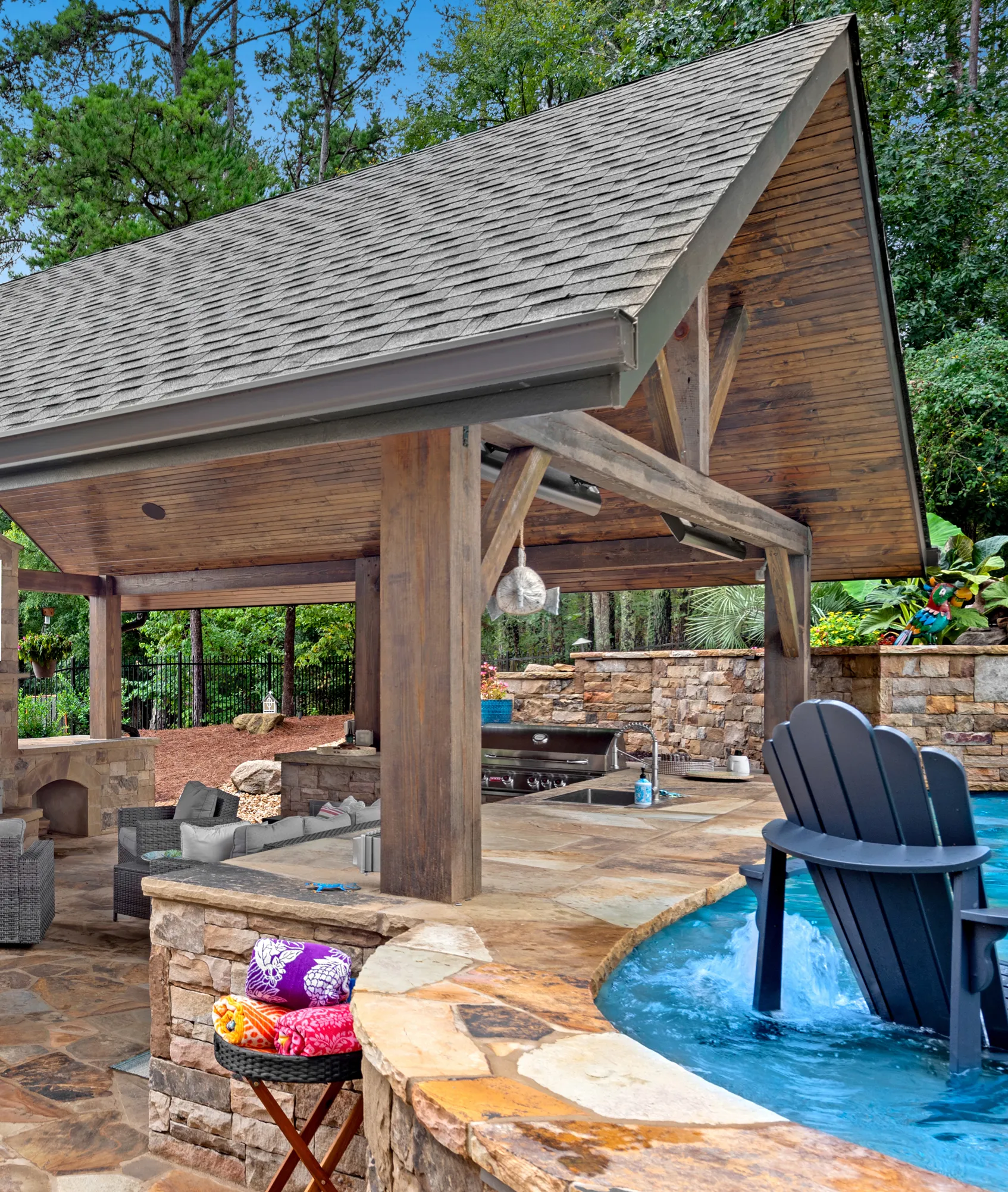 Outdoor living area with a covered stone patio, built-in grill, seating area, and a pool featuring an in-water Adirondack chair. Rolled towels sit on a side table, and the space is surrounded by lush landscaping and trees.