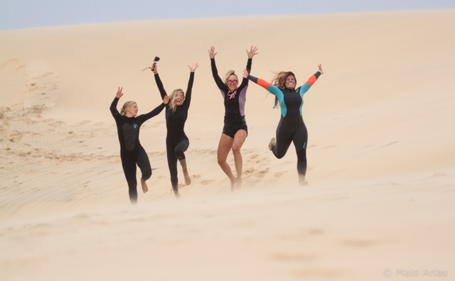 Four women in wetsuits leaping down a sand dune