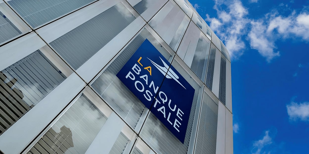 Façade vitrée d'un bâtiment avec le logo bleu de La Banque Postale sur fond de ciel bleu avec quelques nuages.