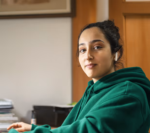 Young woman wearing a dark green hoodie and wireless earbuds, looking confidently at the camera.