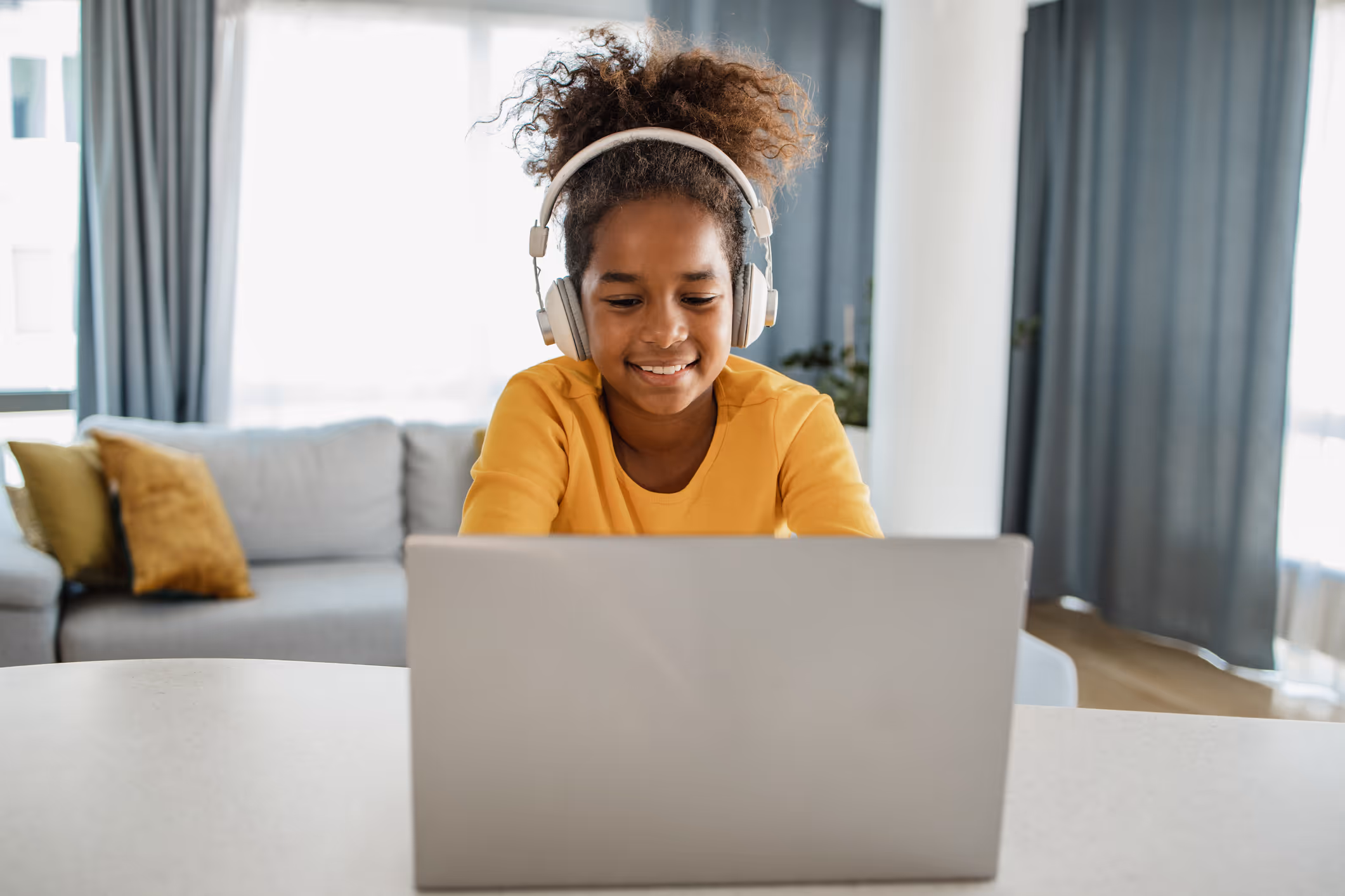 Smiling girl with headphones using laptop at a table in a bright living room.