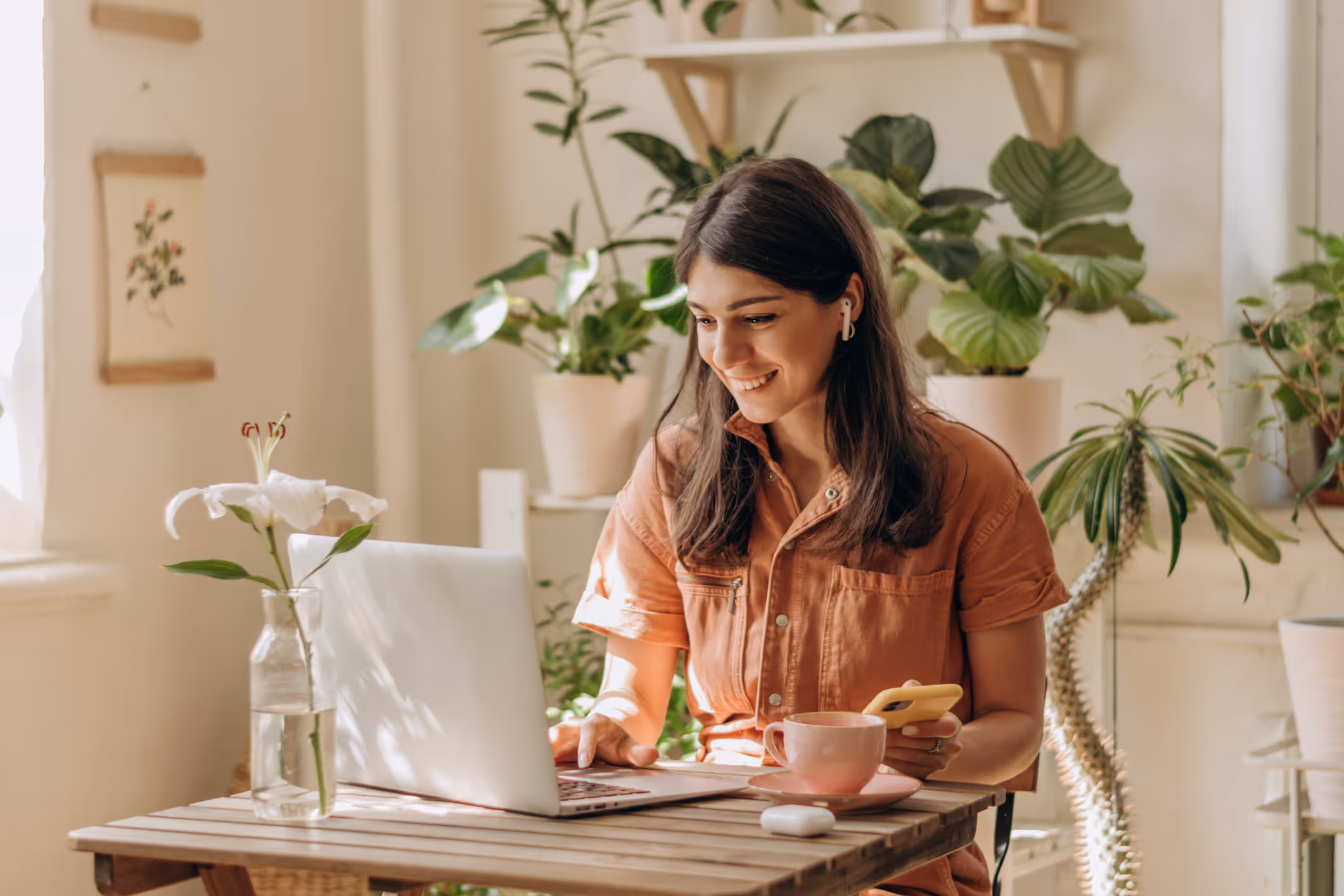 Woman with earbuds smiling while using laptop and holding phone at a table with plants and coffee cup.