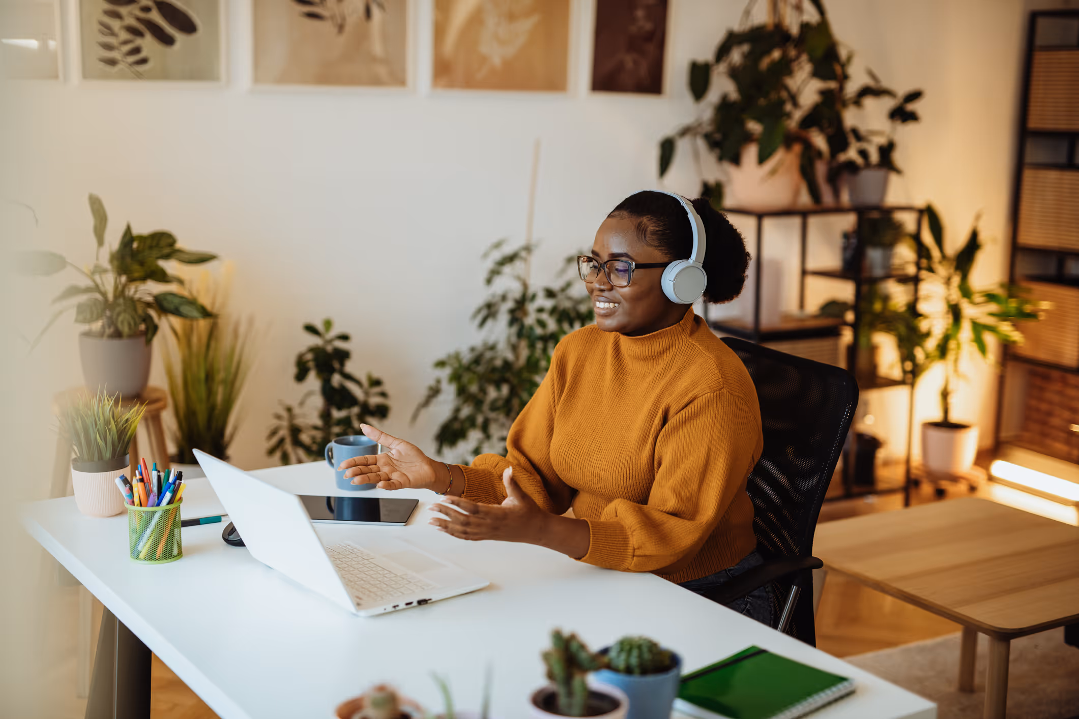 Smiling woman with headphones video chatting at a white desk with plants and stationery.