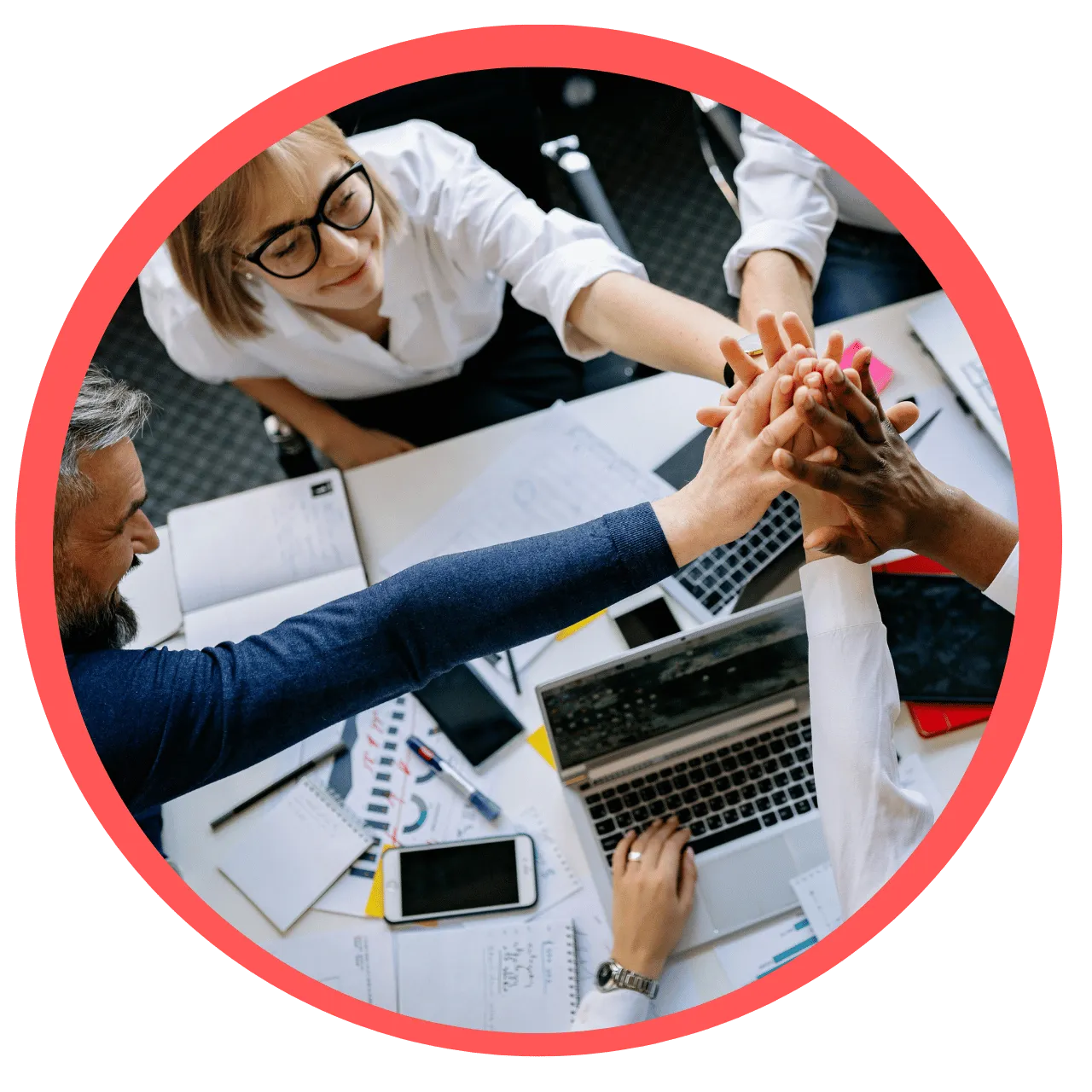 Diverse team of colleagues joining hands in a group high-five over a cluttered office desk with laptops and documents.