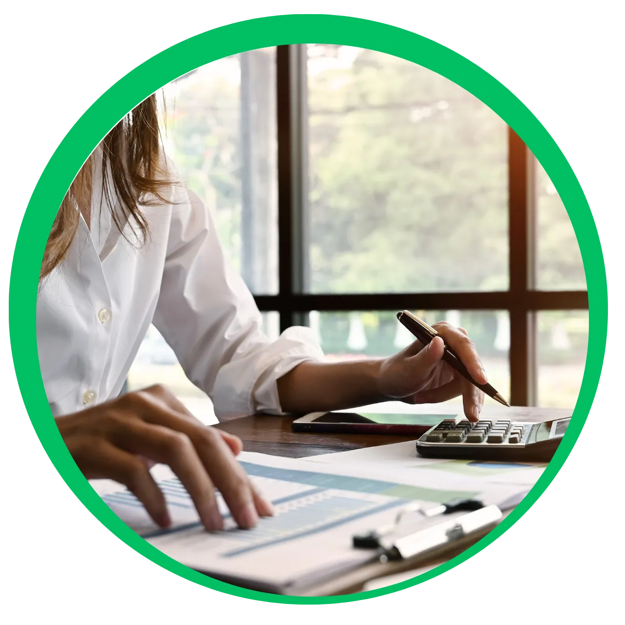 Person in white shirt using a calculator and reviewing documents at a desk by a window.
