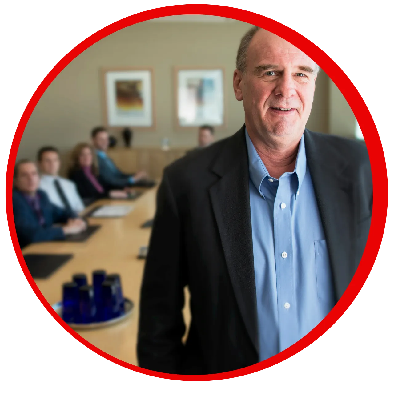 Middle-aged man in a blue shirt and black blazer standing in front of a conference table with five seated people in a meeting room.