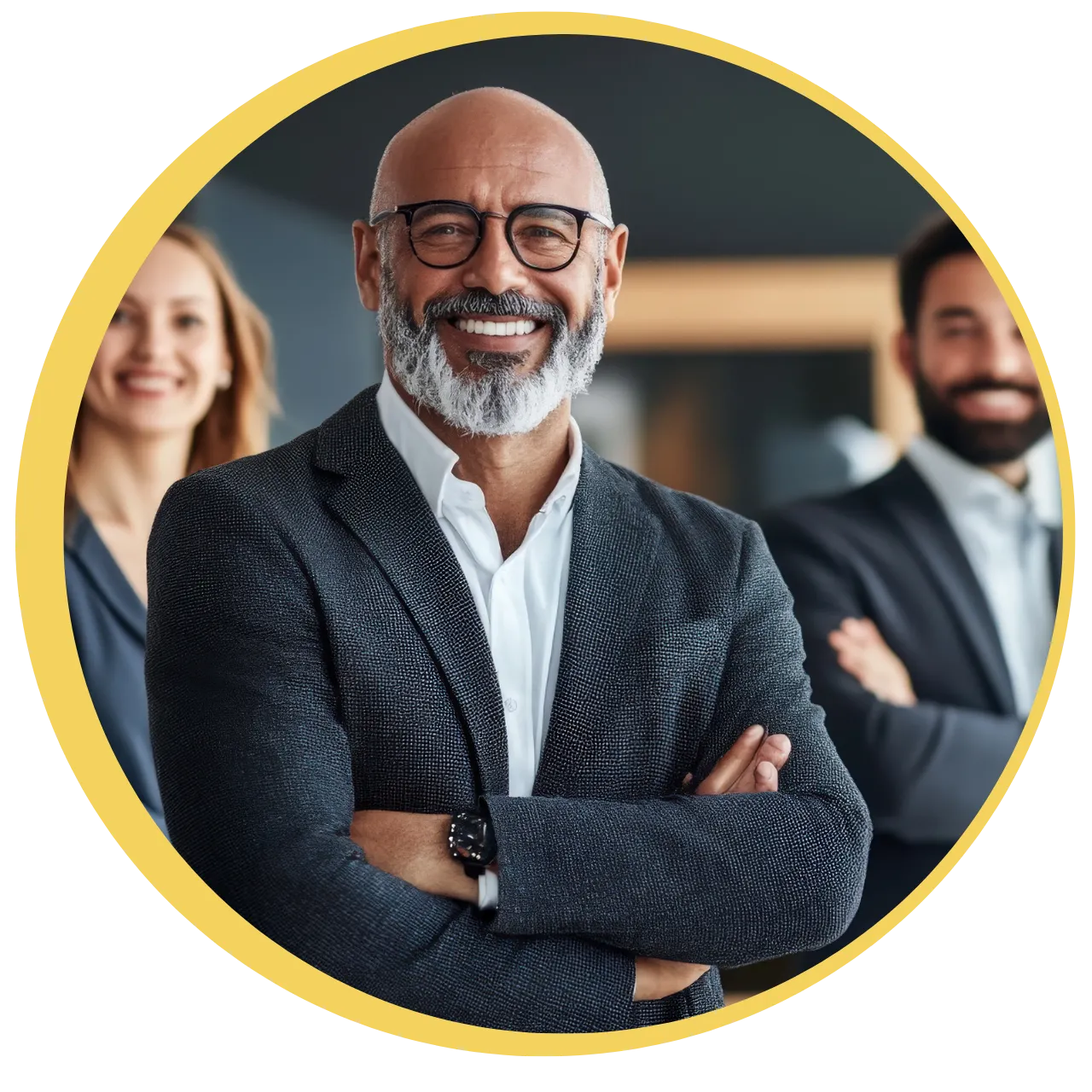 Smiling confident middle-aged man with glasses and gray beard wearing a blazer, standing with crossed arms; two blurred colleagues in the background.