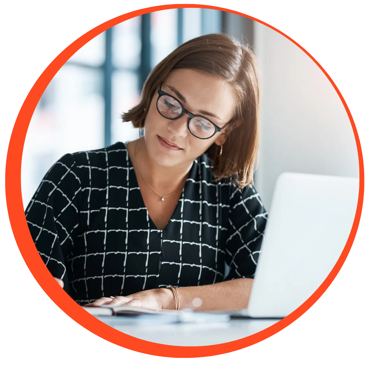 Woman with glasses and short hair writing in a notebook at a desk with a laptop.