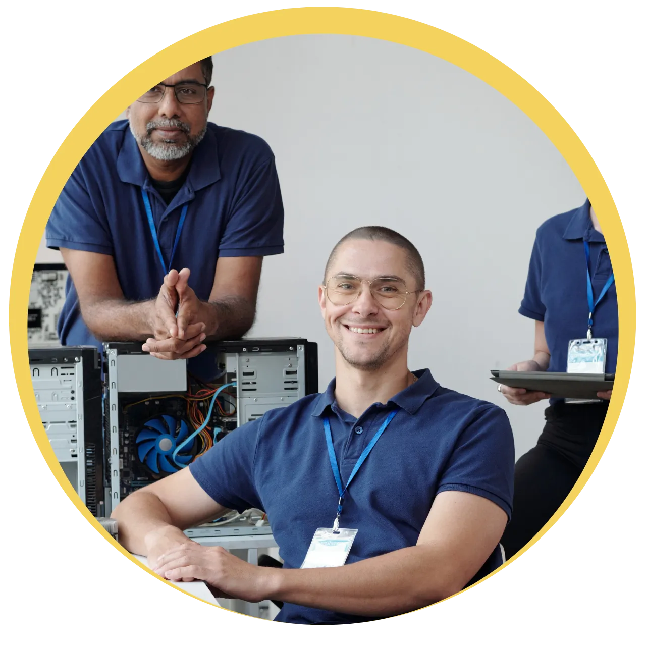Three IT professionals in blue shirts with ID badges, one sitting and smiling, another leaning on a desk with computer hardware, and the third standing holding a tablet.