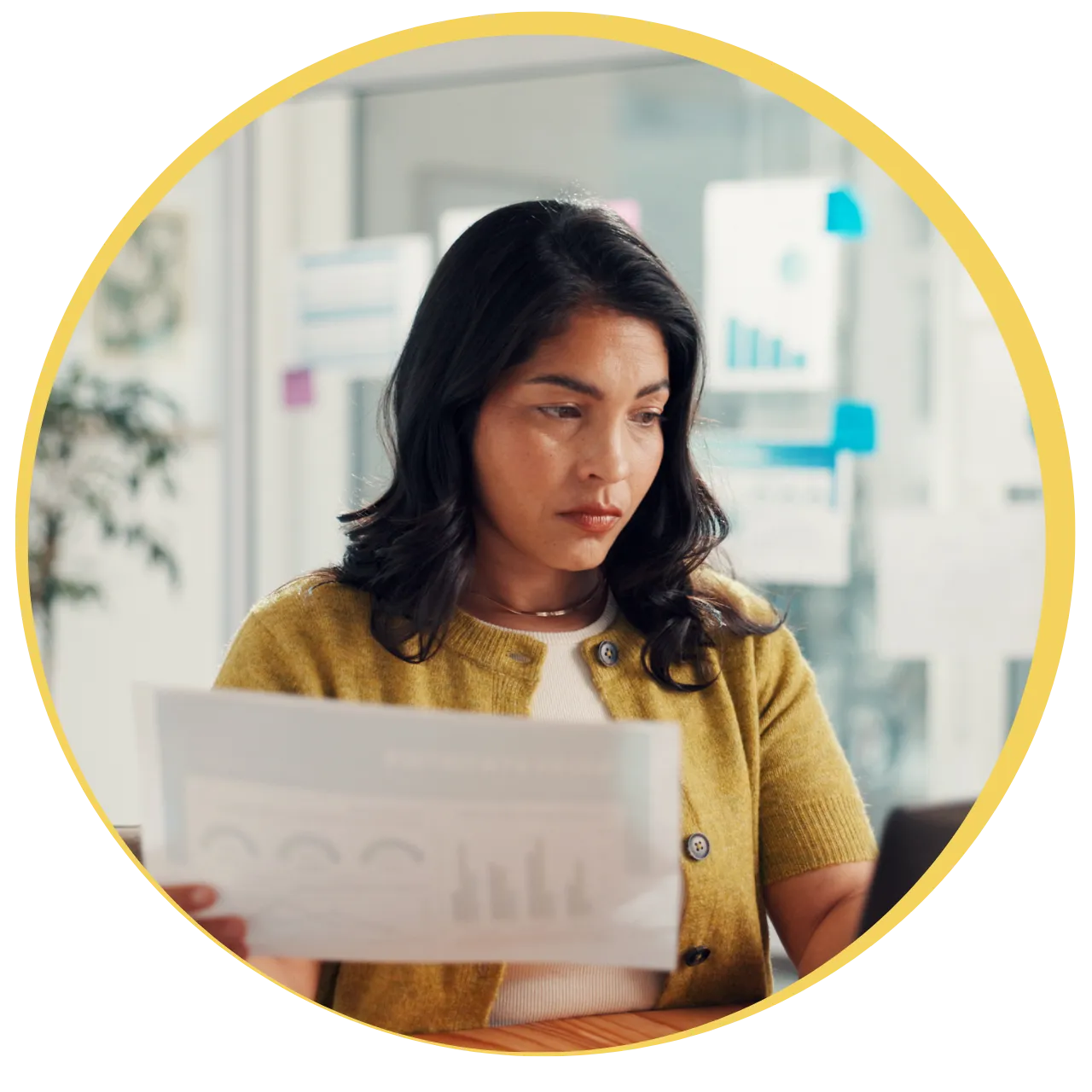 Woman with dark hair wearing a yellow cardigan, reviewing printed charts while working in a bright office.