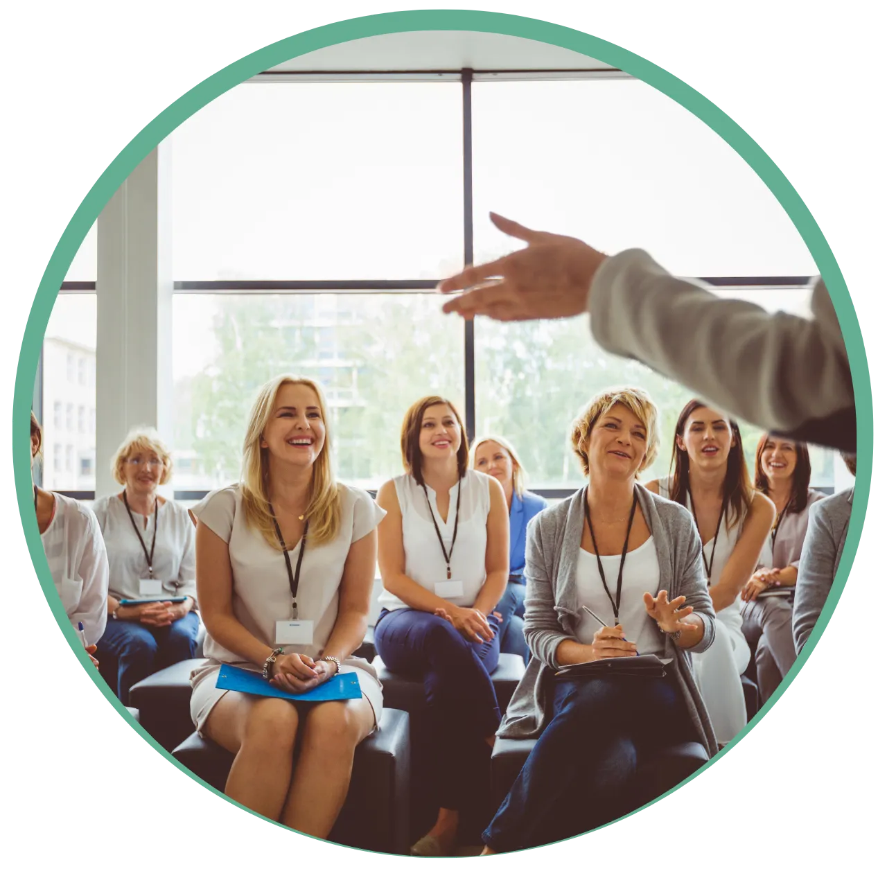 Group of women sitting and smiling attentively during an indoor training or seminar with a speaker gesturing in the foreground.