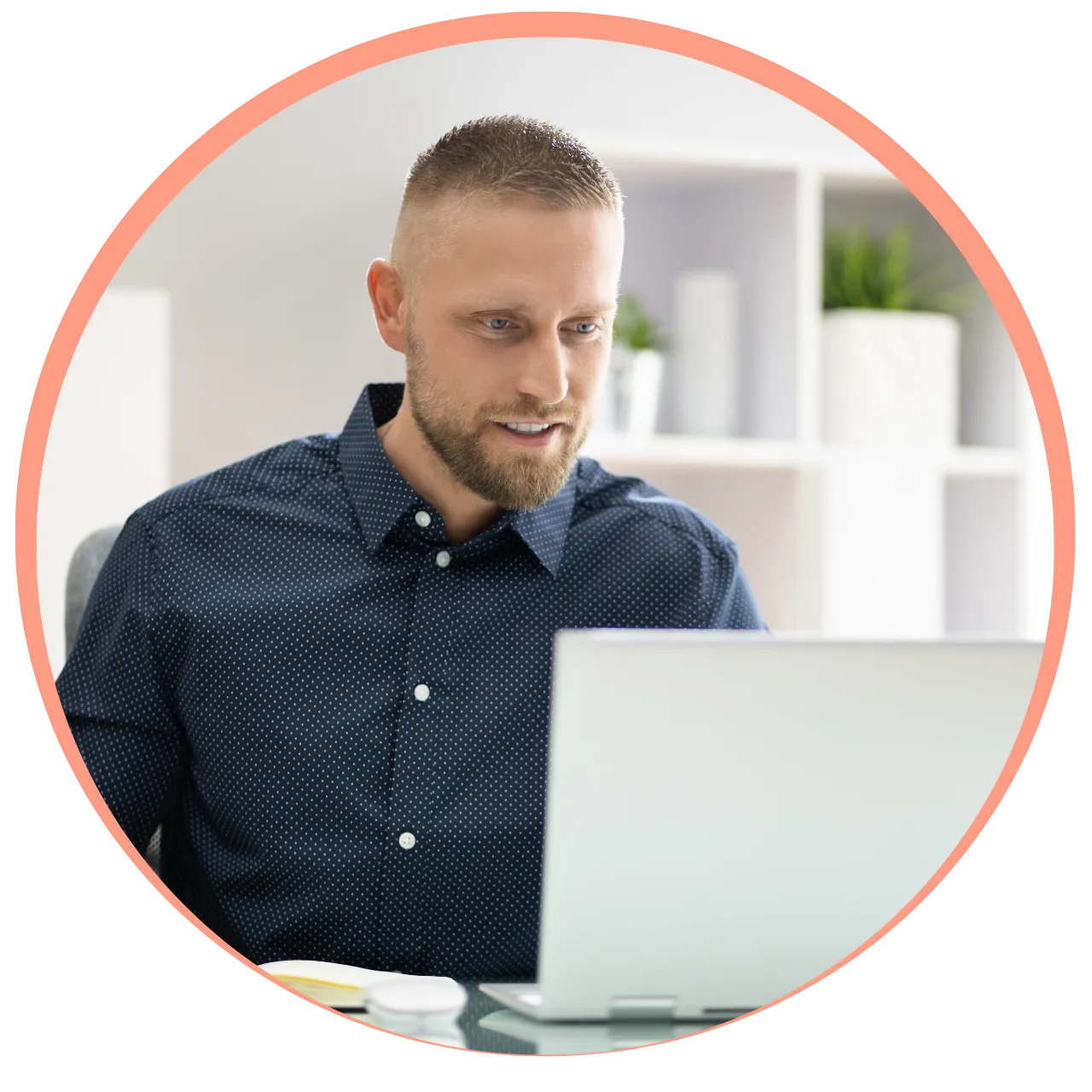 Man with short hair and beard wearing a dark blue patterned shirt working on a laptop in a modern office.