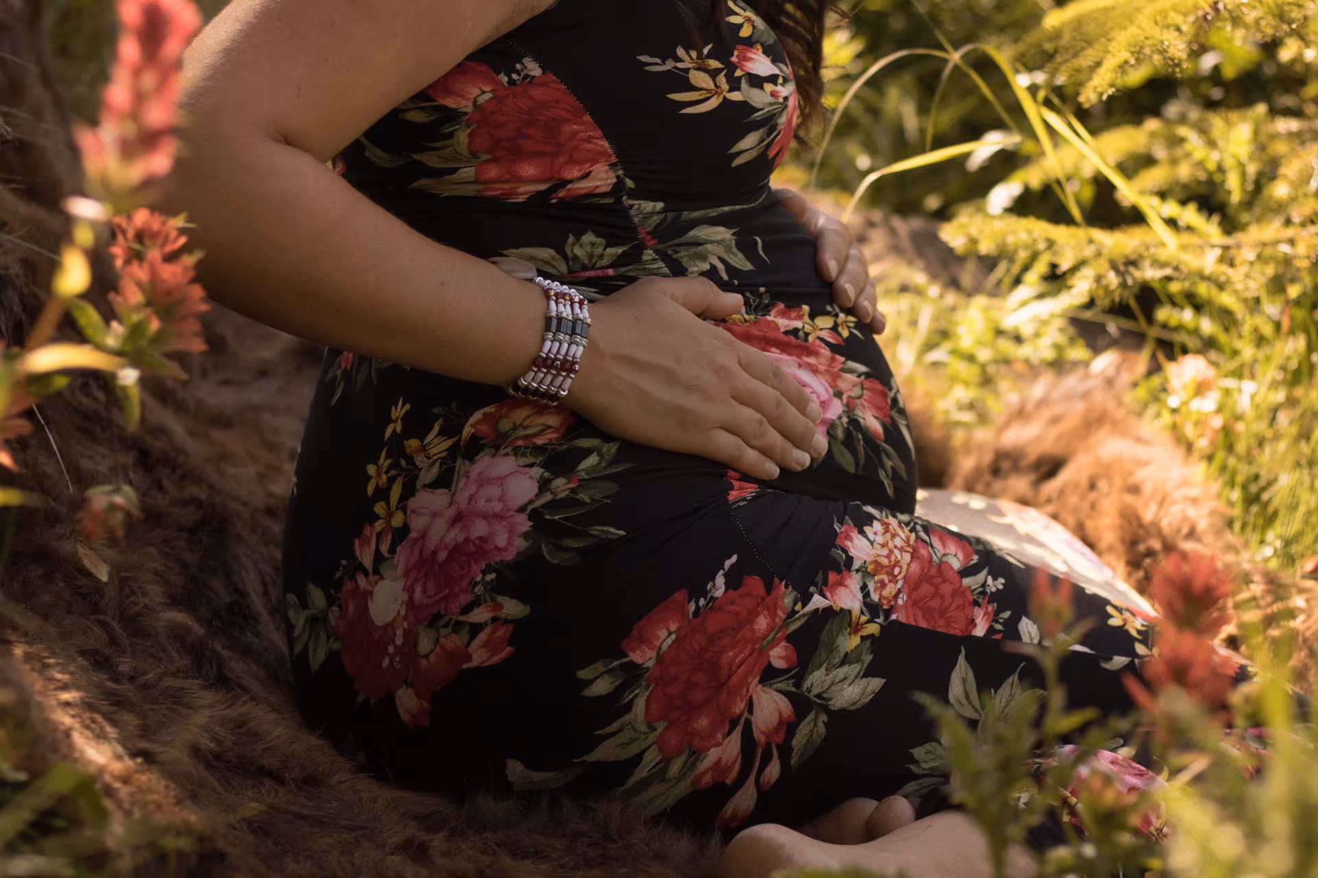 Pregnant indigenous woman sitting outdoors in a floral dress cradling her belly with an eagle feather nearby