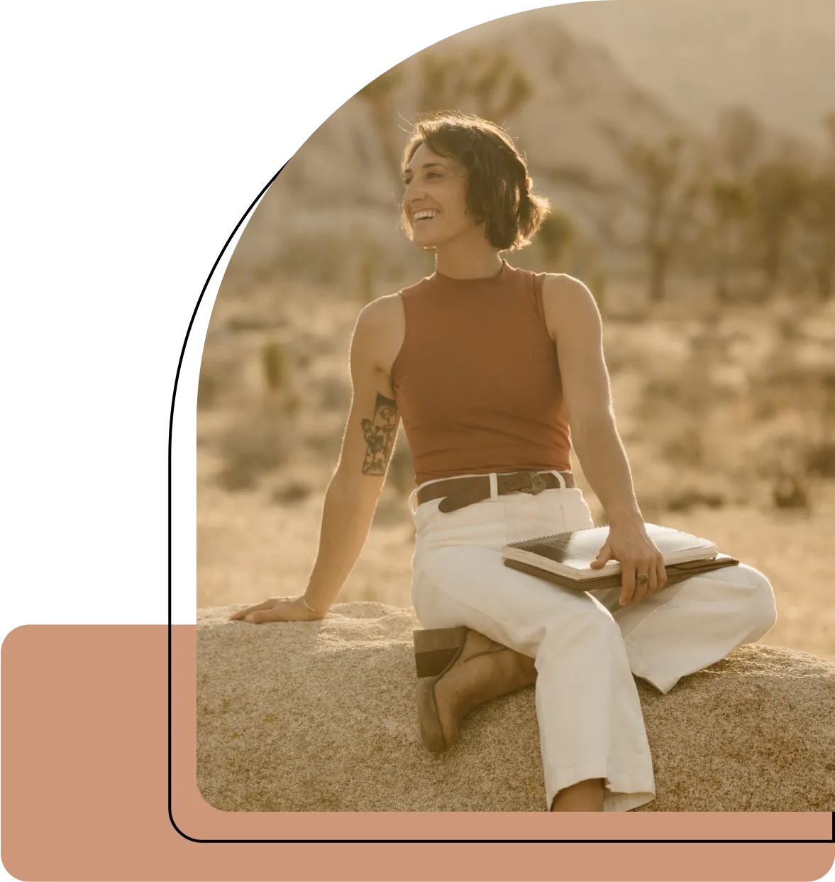 Smiling woman in a sleeveless rust-colored top and white pants sitting on a large rock outdoors, holding notebooks with a desert landscape in the background.