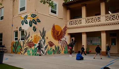 Children playing basketball on a court beside a building with a colorful nature-themed mural featuring birds and plants.