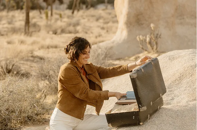 Woman smiling while opening a vintage black suitcase outdoors on a large rock in a desert landscape.
