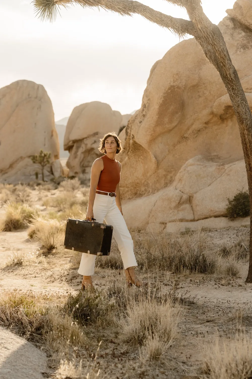 Woman in white pants and sleeveless rust top carrying a vintage suitcase walking in a rocky desert landscape.