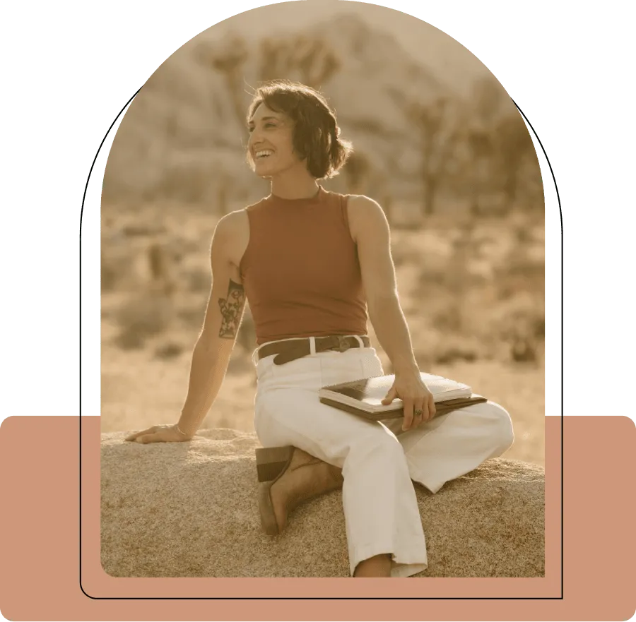 Smiling woman with short hair and a tattoo on her arm sitting on a rock outdoors, holding a notebook.
