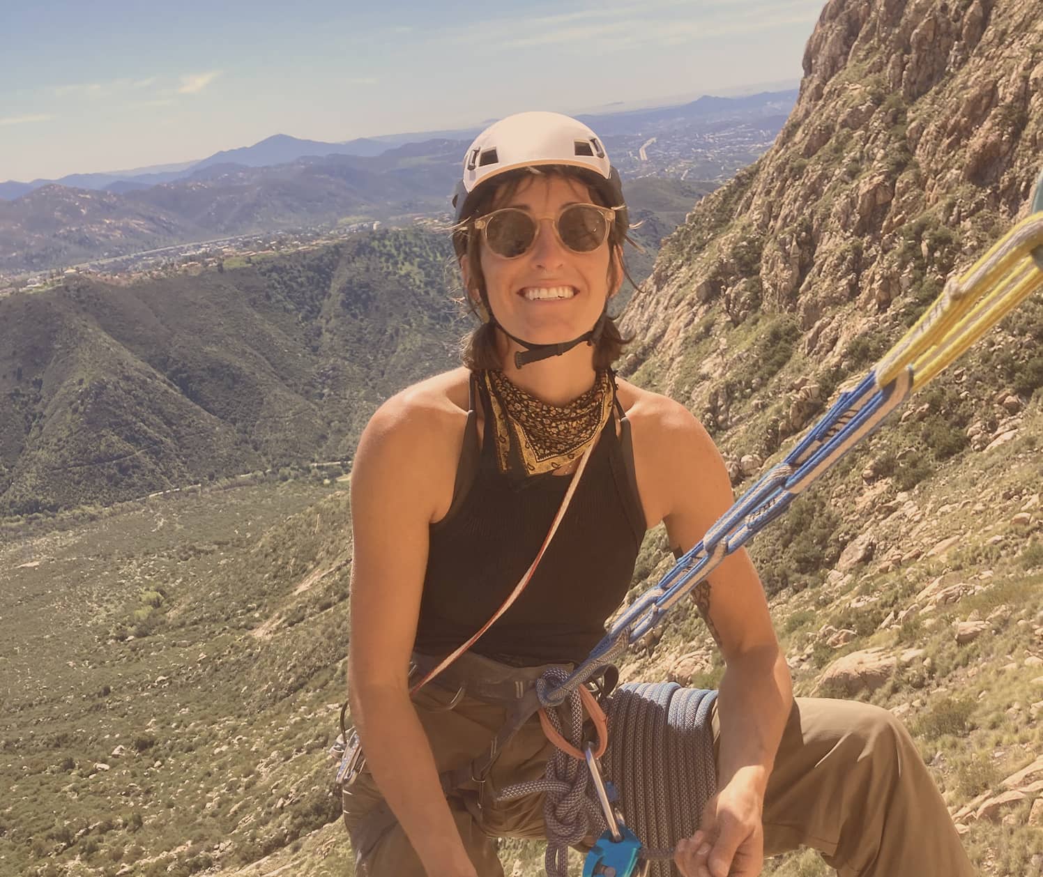 Smiling woman wearing a climbing helmet and sunglasses, secured with ropes on a rocky mountain with a scenic valley in the background.