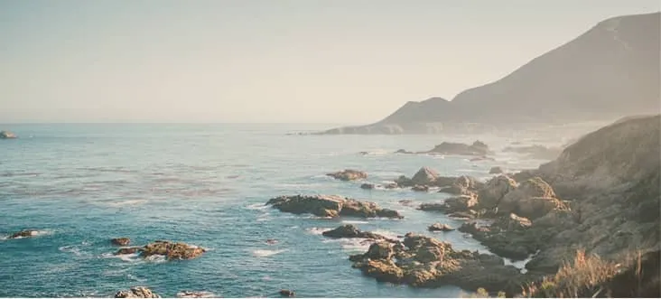 Rocky coastline with calm ocean waves and misty mountains in the background under a clear sky.