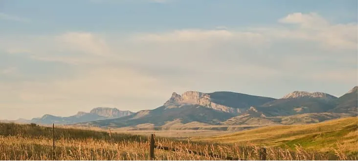 Grassy field with wooden fence posts and a mountain range under a partly cloudy sky.