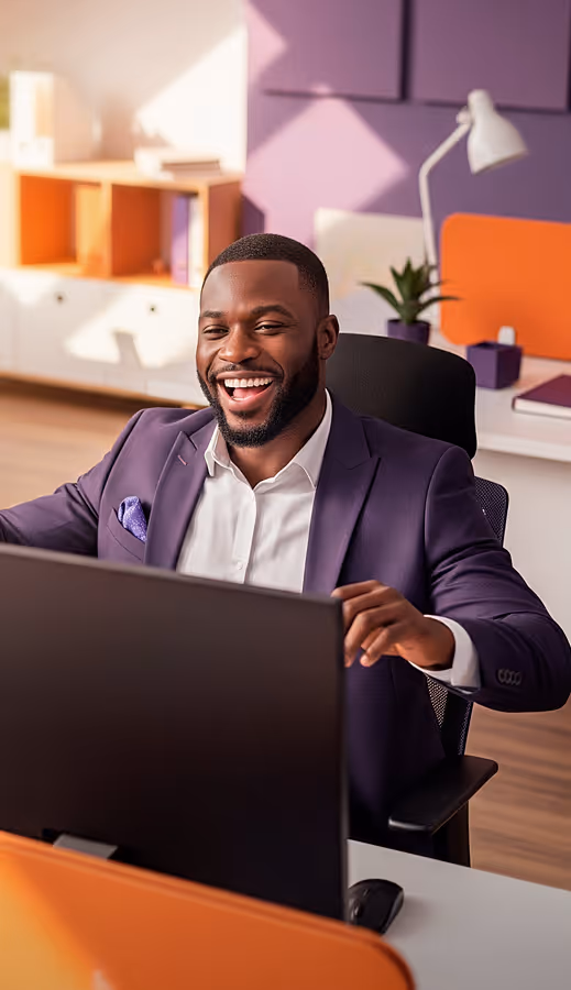Homem sorridente, vestido com terno roxo e camisa branca, sentado em frente a um computador em um escritório moderno.