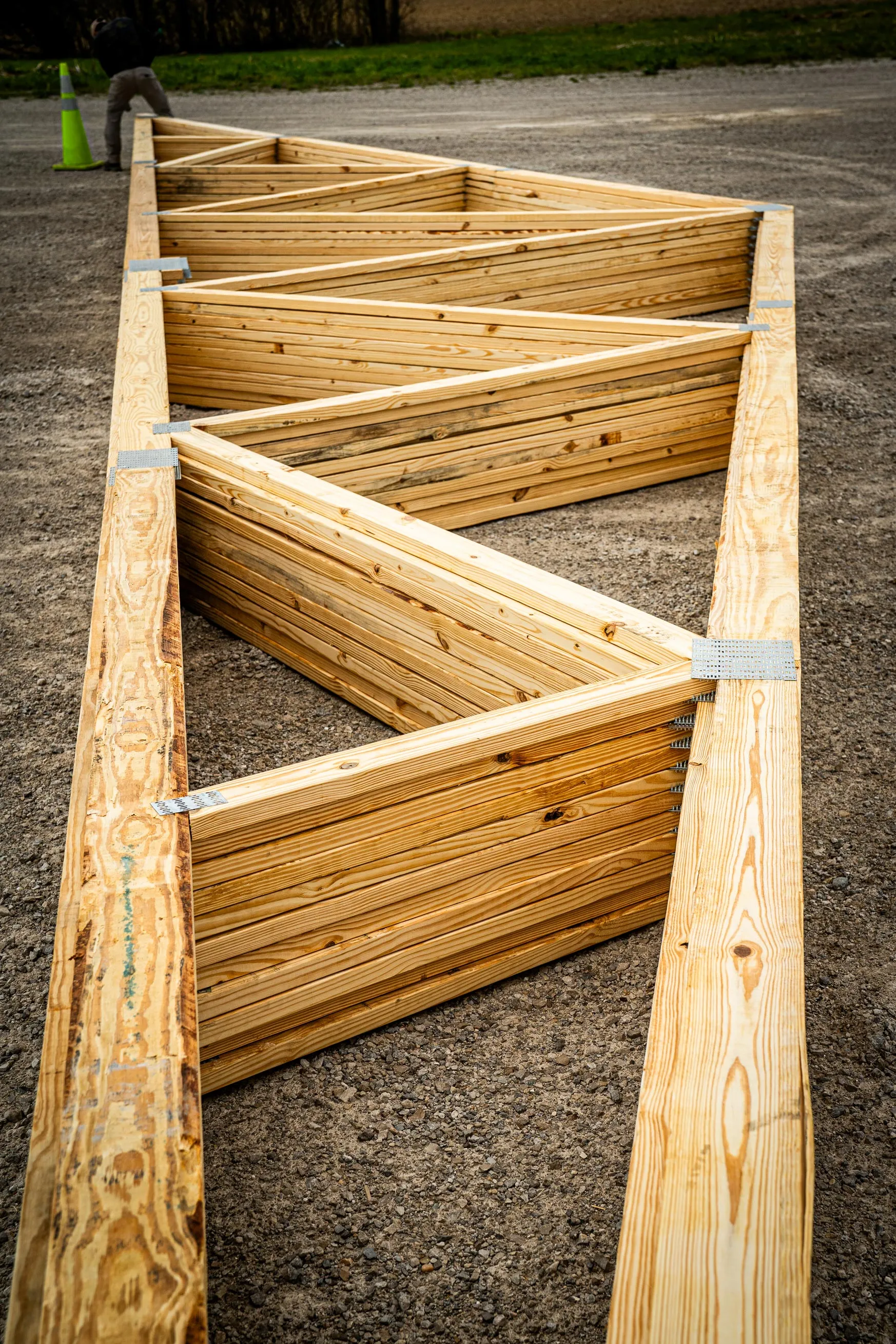 Large wooden roof truss with diagonal supports lying on gravel ground at a construction site.