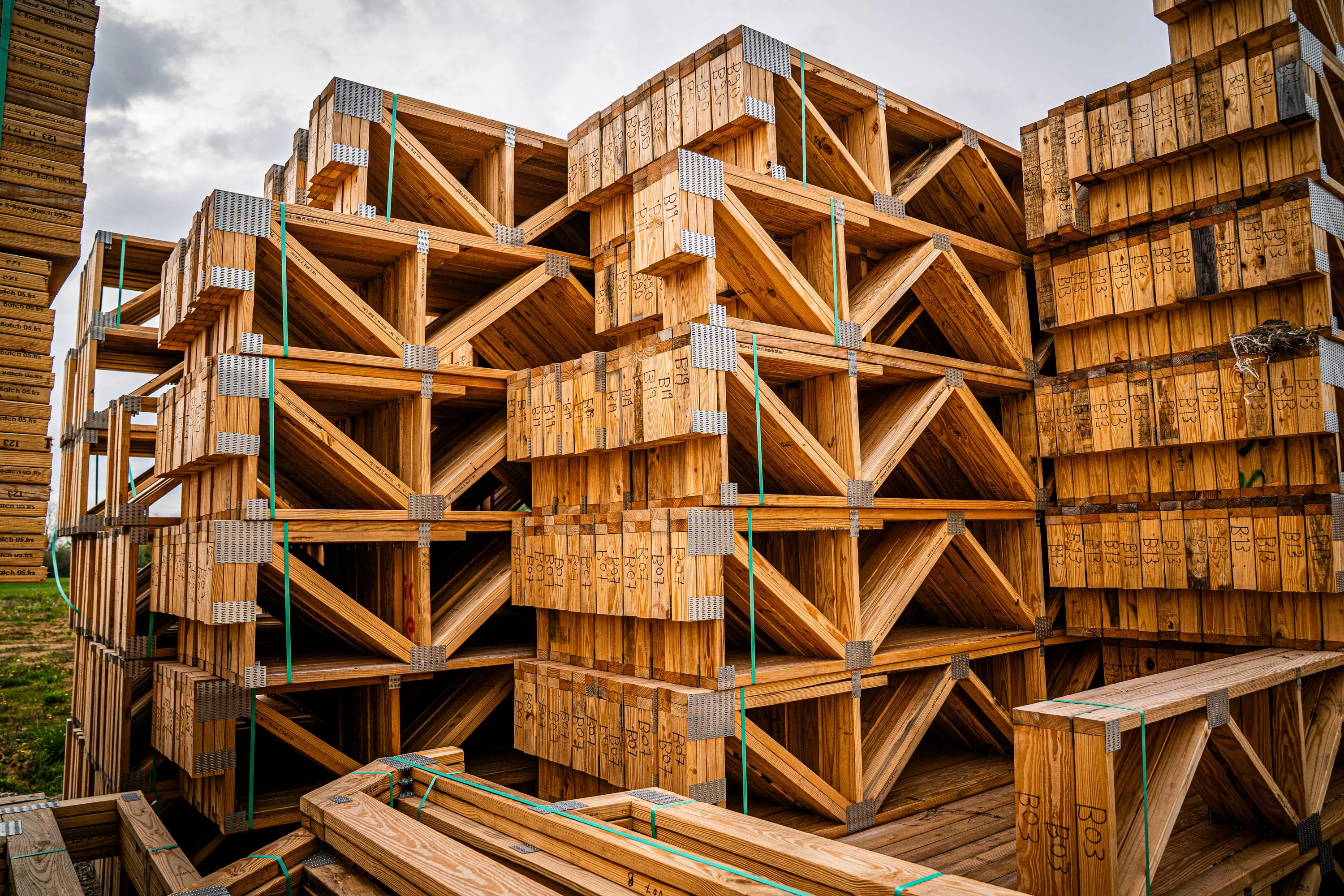 Stacks of large wooden floor trusses bound with green straps arranged outdoors under a cloudy sky.