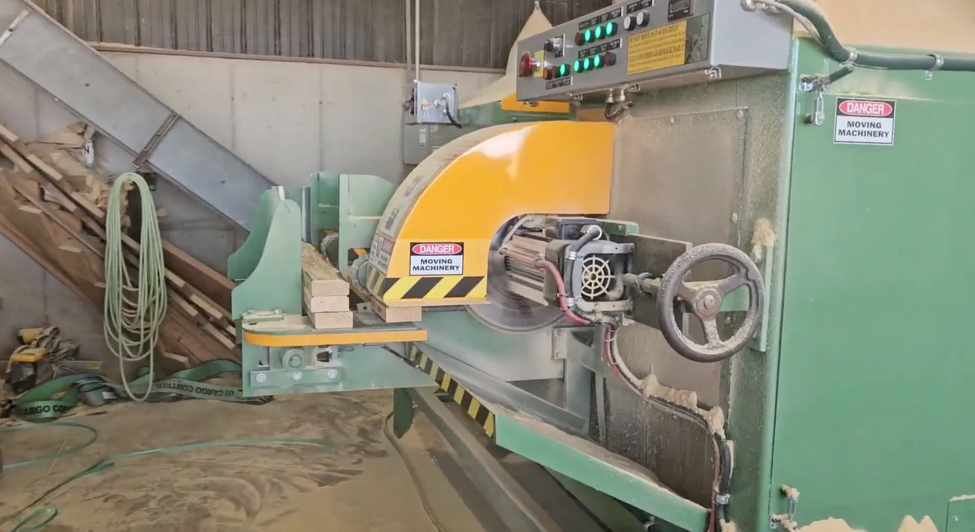 Industrial green and yellow saw machine cutting wooden planks in a workshop with warning labels.