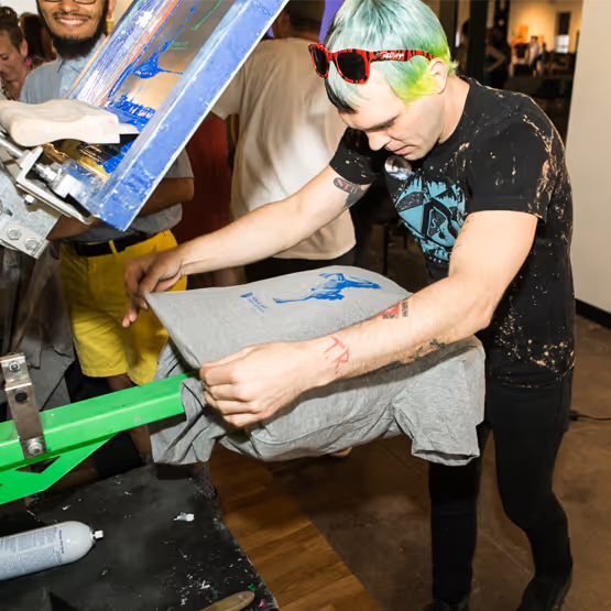 Person with dyed green hair screen printing a t-shirt at a live Denver Public Art event.