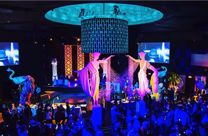 Vibrant event scene with dramatic lighting, tall costumed performers on pedestals, and a crowd gathered in a large venue.