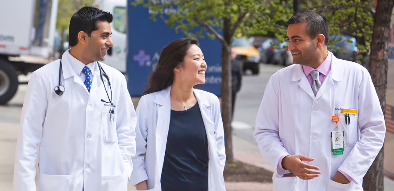 Three smiling medical professionals in white coats walking and talking on a city sidewalk.
