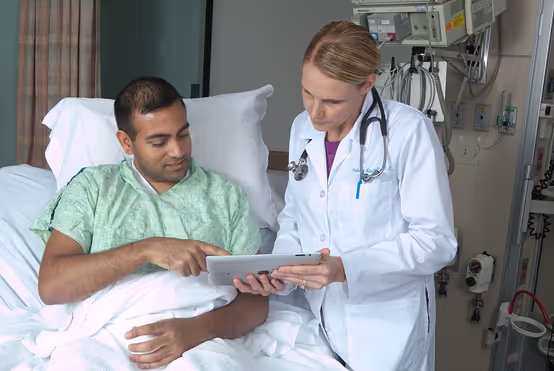 Physician in a white coat showing information on a tablet to a patient sitting in a hospital bed.